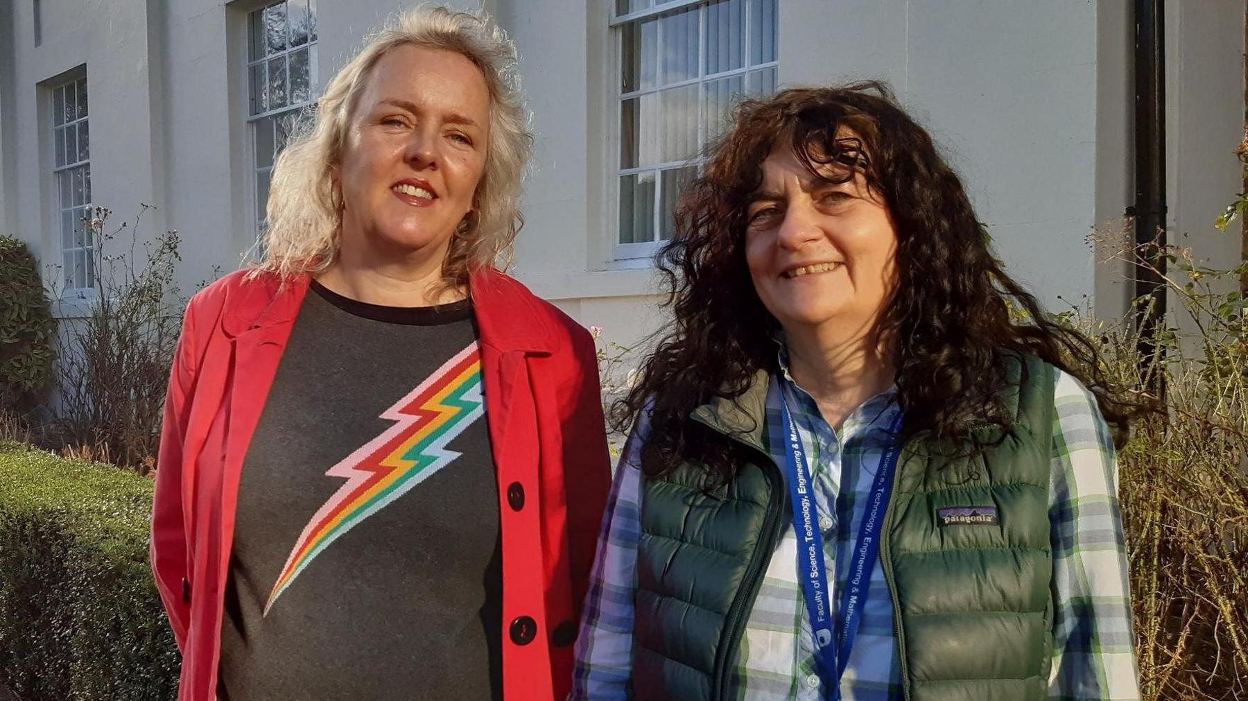 Prof Catriona Havard and Dr Hayley Ryder stand side-by-side outside a white building with sash windows, with hedges in the background. Prof Havard is wearing a red jacket, open, with two black buttons, over a grey t-shirt with a rainbow coloured lightning strike. She is white with blond shoulder length hair. Dr Ryder has curly long dark brown hair and is wearing a checked shirt with a green gilet over it, and an OU blue lanyard around her neck.