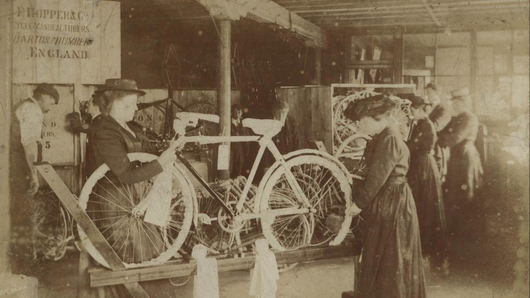 A black and white archive photo of women wearing long dresses and hats with their hair tied back as they hold a bicycle which is propped up on a stand. They are working on it. Behind them are other women working, while a man is on the far left.