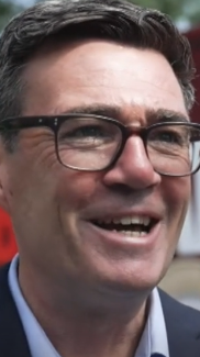 Andy Burnham smiles during an interview. He is a middle aged man with black hair, and wears a light blue shirt with a navy suit jacket and black square-framed glasses. 