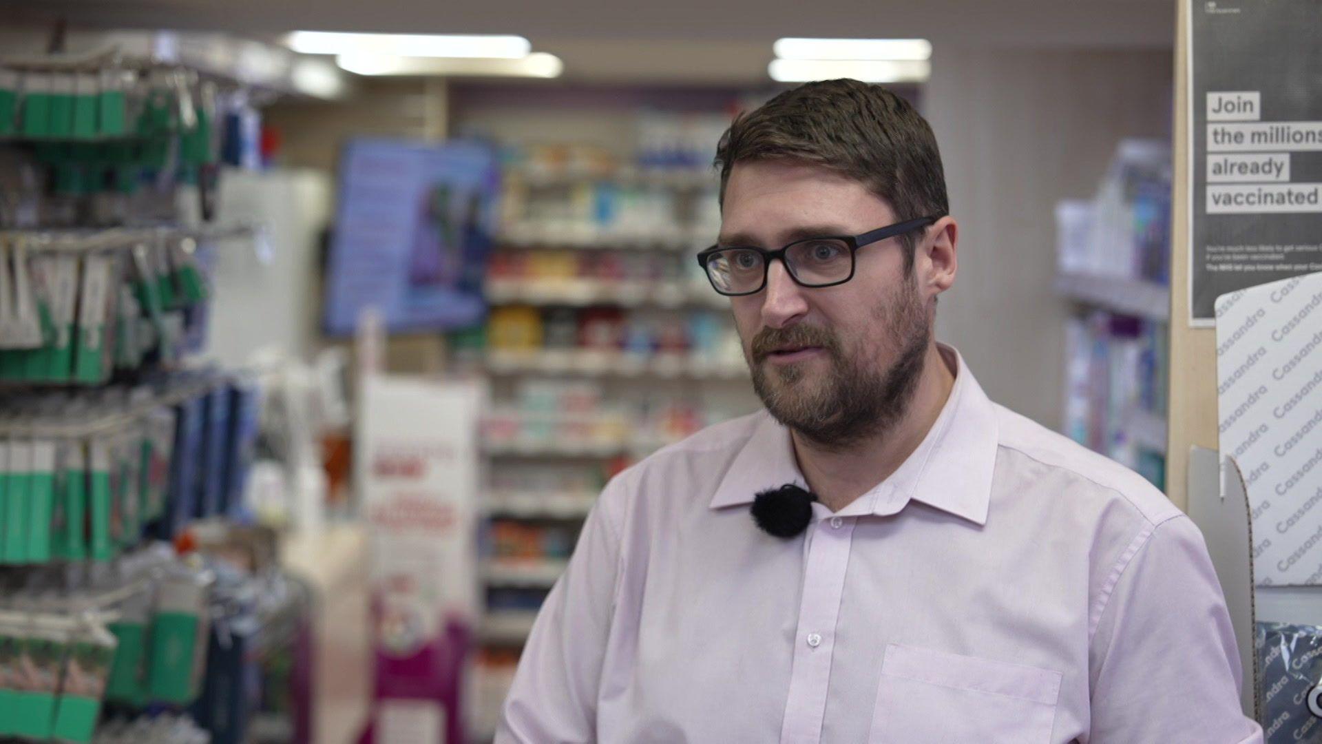The image shows the inside of a pharmacy or chemist shop. There are shelves stocked with various health and personal care products on the left and in the background. A person wearing a light-coloured shirt with a clip-on microphone is standing in the foreground. On the right side, there is a poster that reads: “Join the millions already vaccinated”.