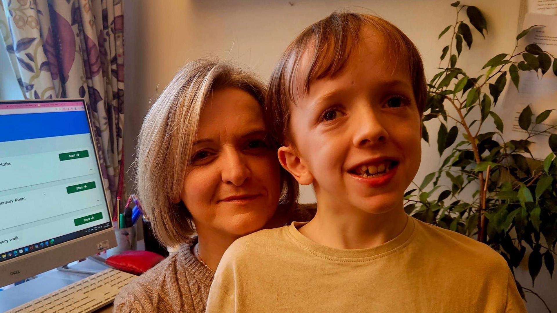 Wendy Gallagher and her son Andrew smiling at the camera. Andrew is slightly in front of Wendy. He has fair hair with a fringe and is wearing a yellow top. Wendy is wearing a beige jumper and has shoulder length fair hair. In the background, there is a computer and a green plant.