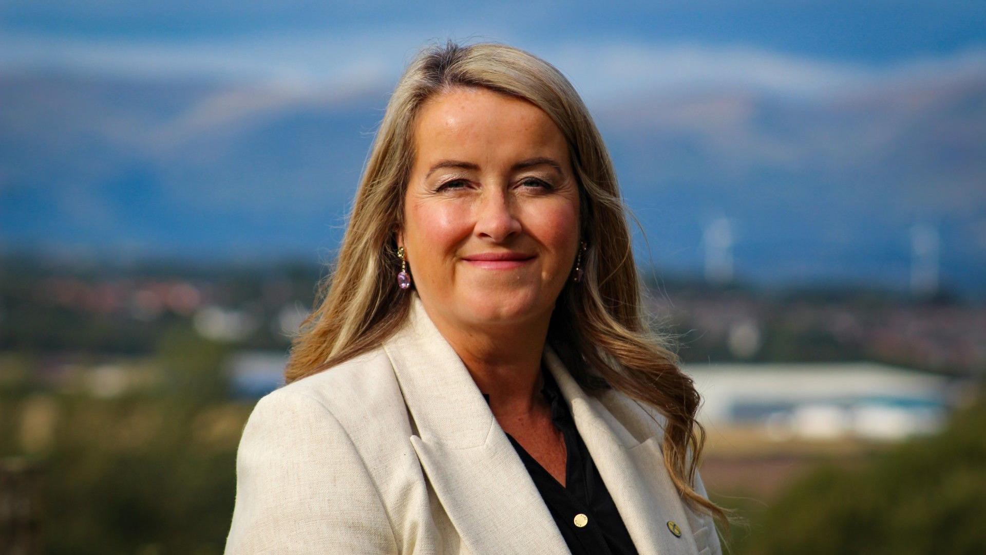 Carol Beattie in a headshot style picture. She is wearing a cream blazer and smiling at the camera. She is standing outside but the background is out of focus. 