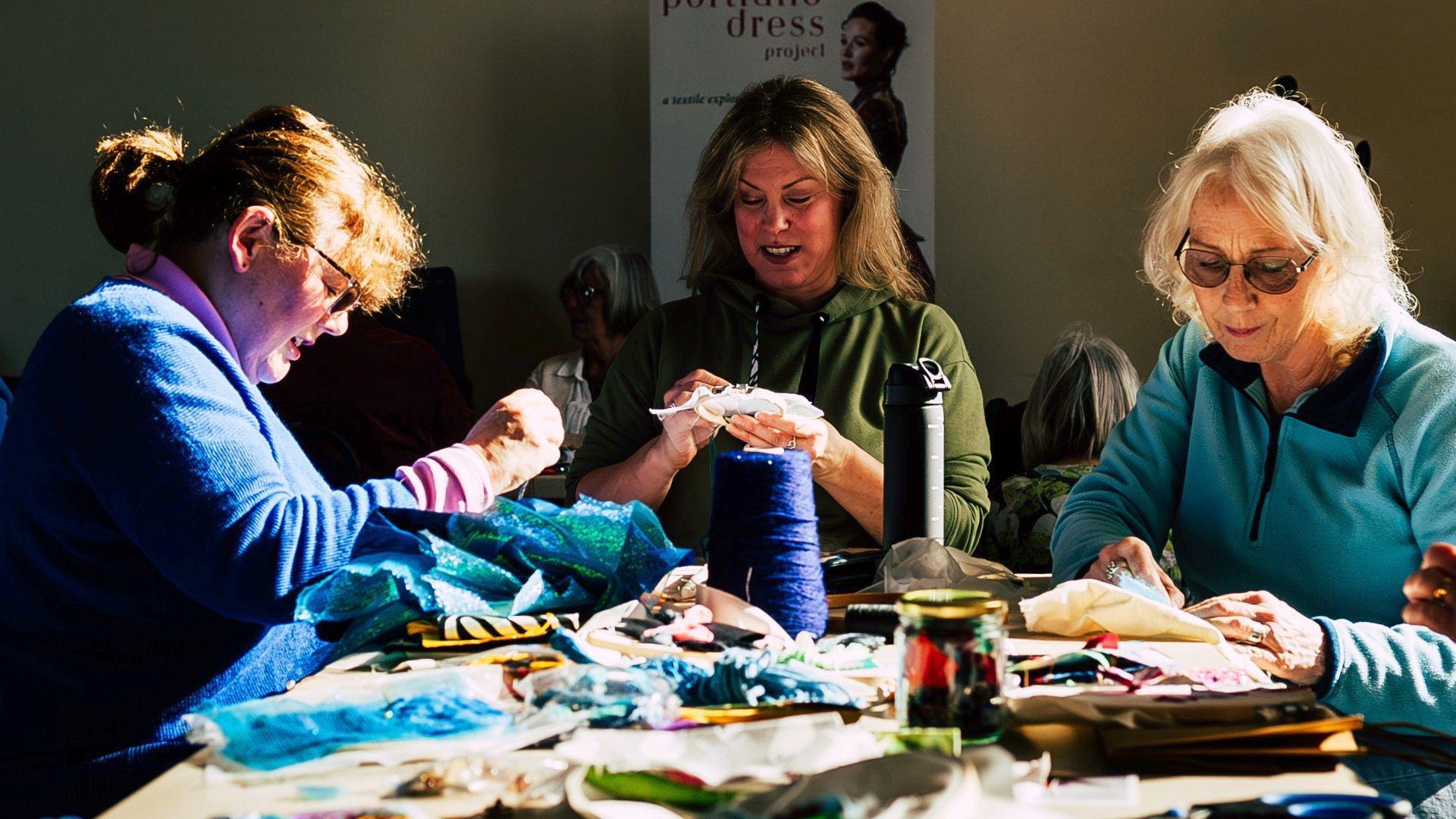 Three women are sat around a table, sewing. There are various sewing materials on the table, pieces of blue fabric, papers, spools of thread. The woman to the left has light brown hair tied back in a ponytail and has a fringe. She is wearing glass, a pink turtleneck long sleeved top with a blue woollen jumper over the top, she is looking down at her work. In the centre, a woman with dark blonde mid-length hair looks down at her work. She is wearing a khaki green hoodie. The woman to the right has white short hair and round framed glasses on. She is wearing a light blue zip up fleece jumper.