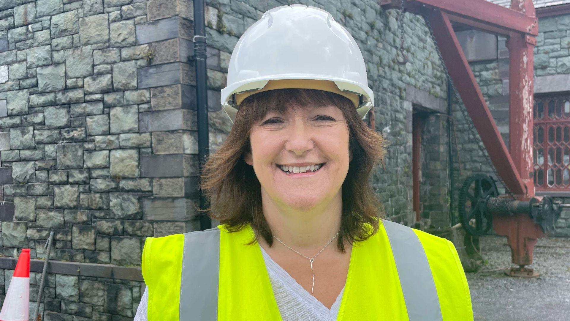 Llun o Elen Roberts yn sefyll ar safle'r amgueddfa. Mae'n gwisgo helmed gwyn a fest 'high-vis'. Mae adeilad cerrig yn y cefndir.