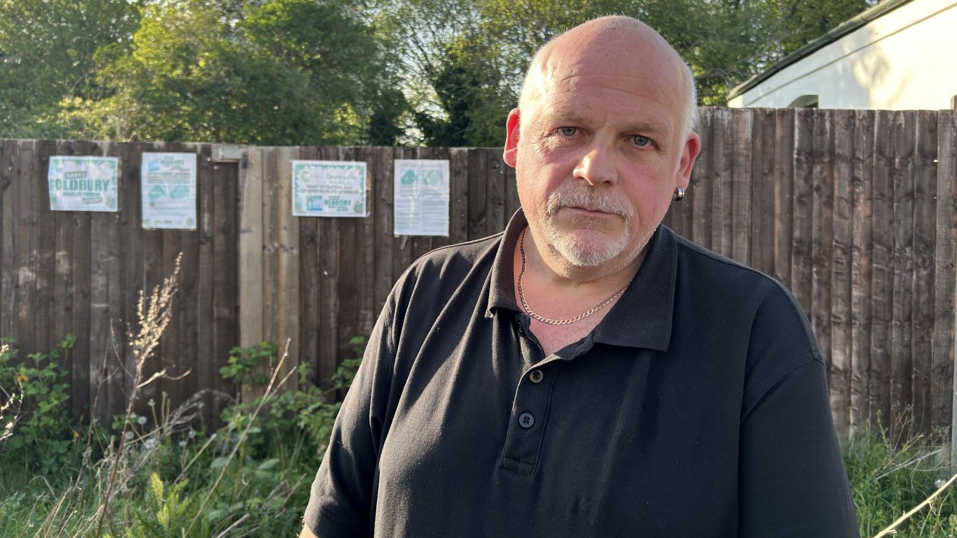 Phil Shakespeare is bald with patches of white hair on each side and a white goatee beard. He is wearing a gold necklace and a black polo shirt. He is standing next to signs pinned on a brown fence opposing the work in Oldbury.