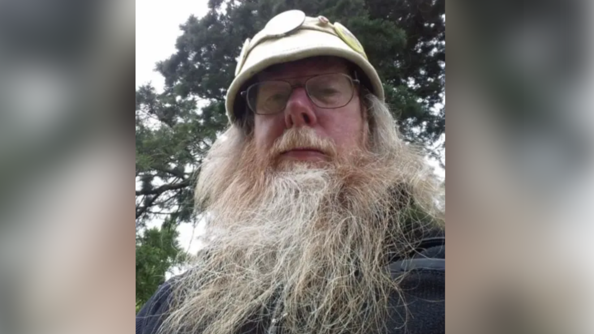 The Baron has a long grey beard and is photographed outside wearing a beige coloured pith helmet