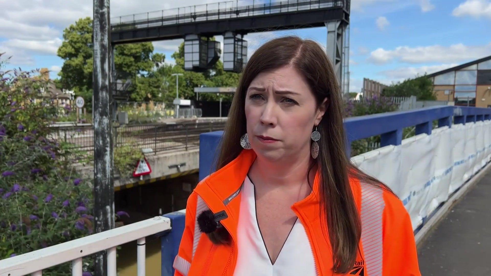 A brown haired woman from the shoulders up, wearing a high-vis jacket. In the background there is a railway bridge