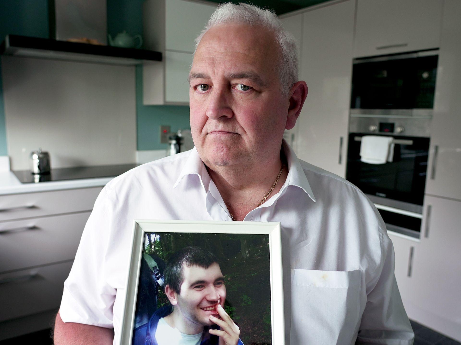Glynn Brown stands in his white kitchen holding a framed photo of his disabled son Aaron. He looks into the camera with a serious expression and the photo of Aaron features him smiling.