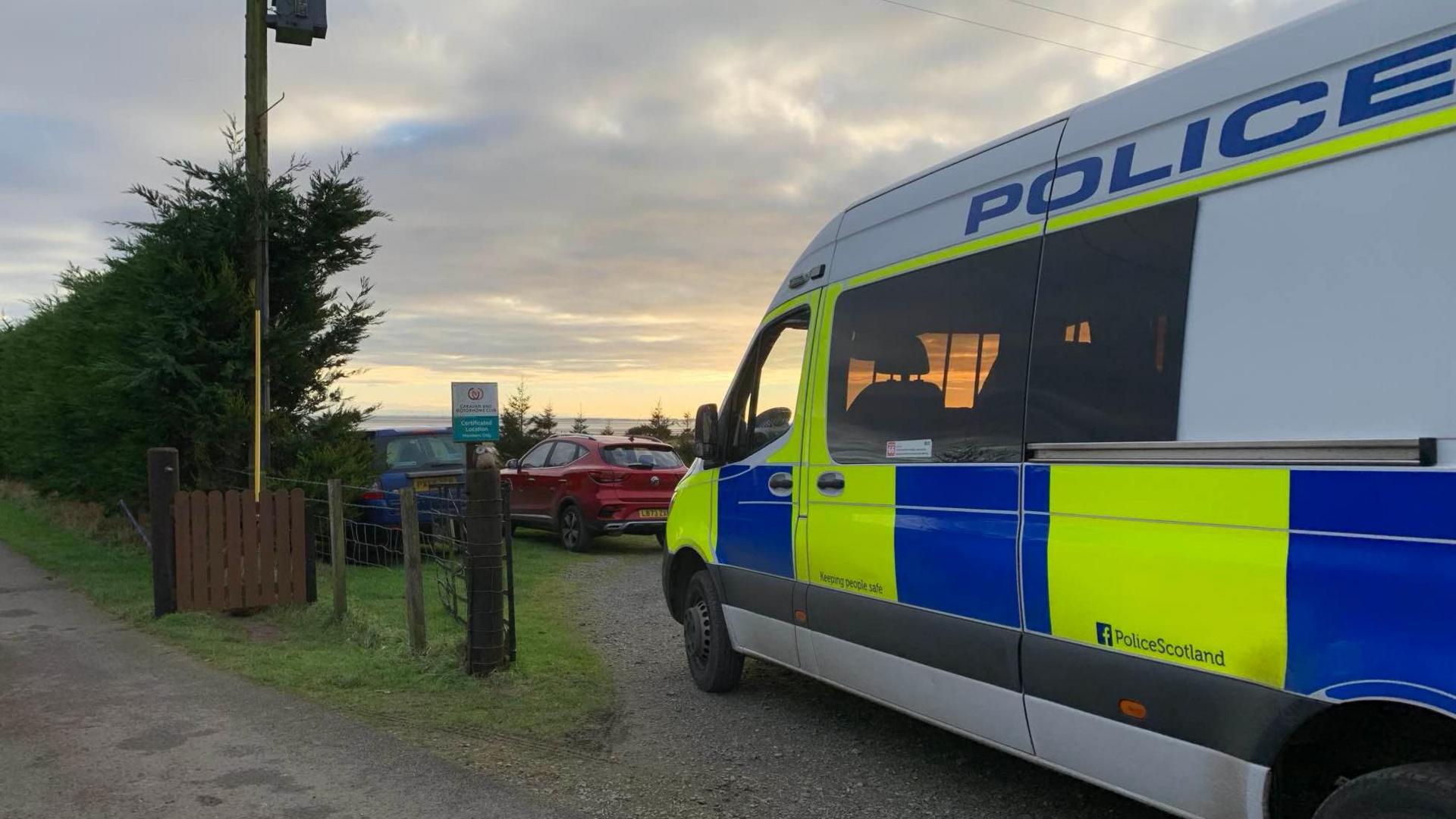 Yellow and blue police van parked on a dirt track behind cars with a hedge on the left. The sky is orange