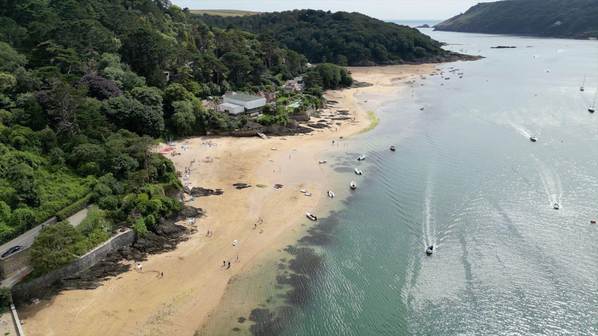 Aerial view of an estuary in the sunshine with people on the beach and yachts in the water