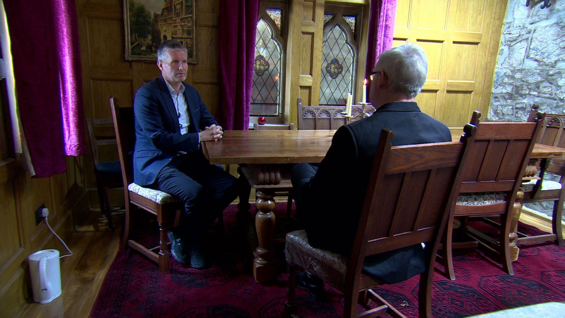 91ȱ reporter Robbie Meredith wearing suit sitting opposite Jim Dowson, whose back is to the camera