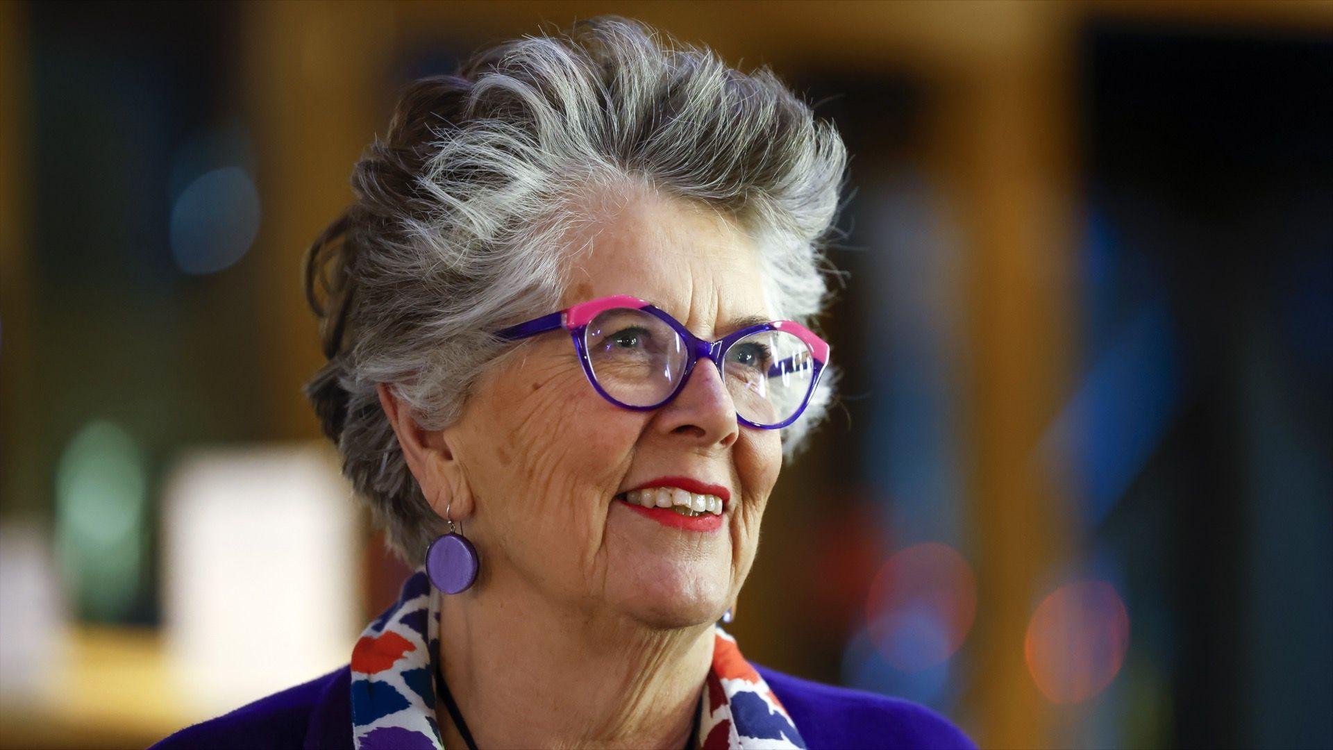 Dame Prue Leith smiling, wearing purple and pink glasses, a patterned scarf and a purple jumper