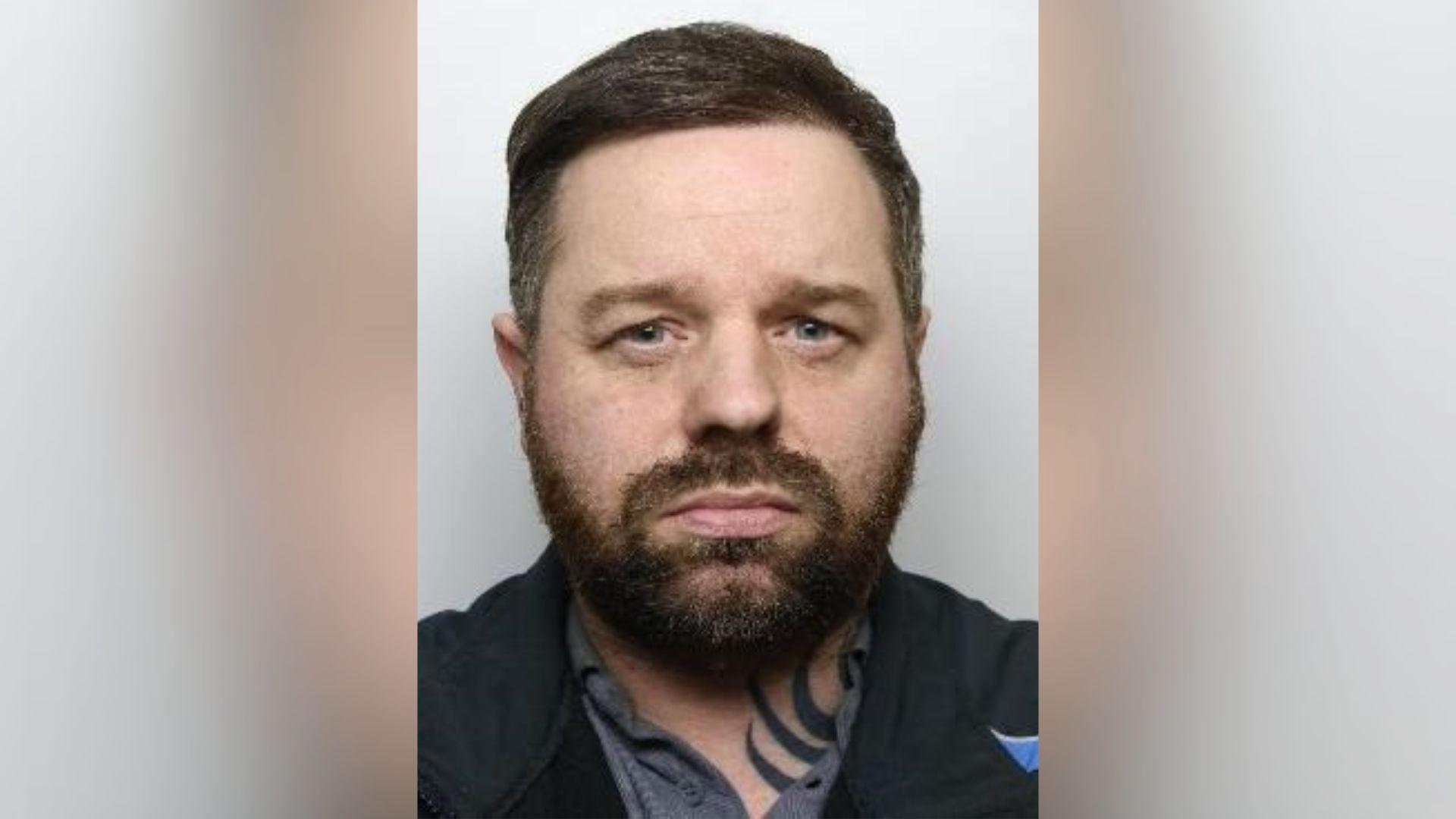 A police mugshot. Webster wears a black tracksuit jacket and a grey polo neck. He has a neck tattoo but it is unclear what of. He has quite a wide face. He has a brown beard and short, brown hair. 
