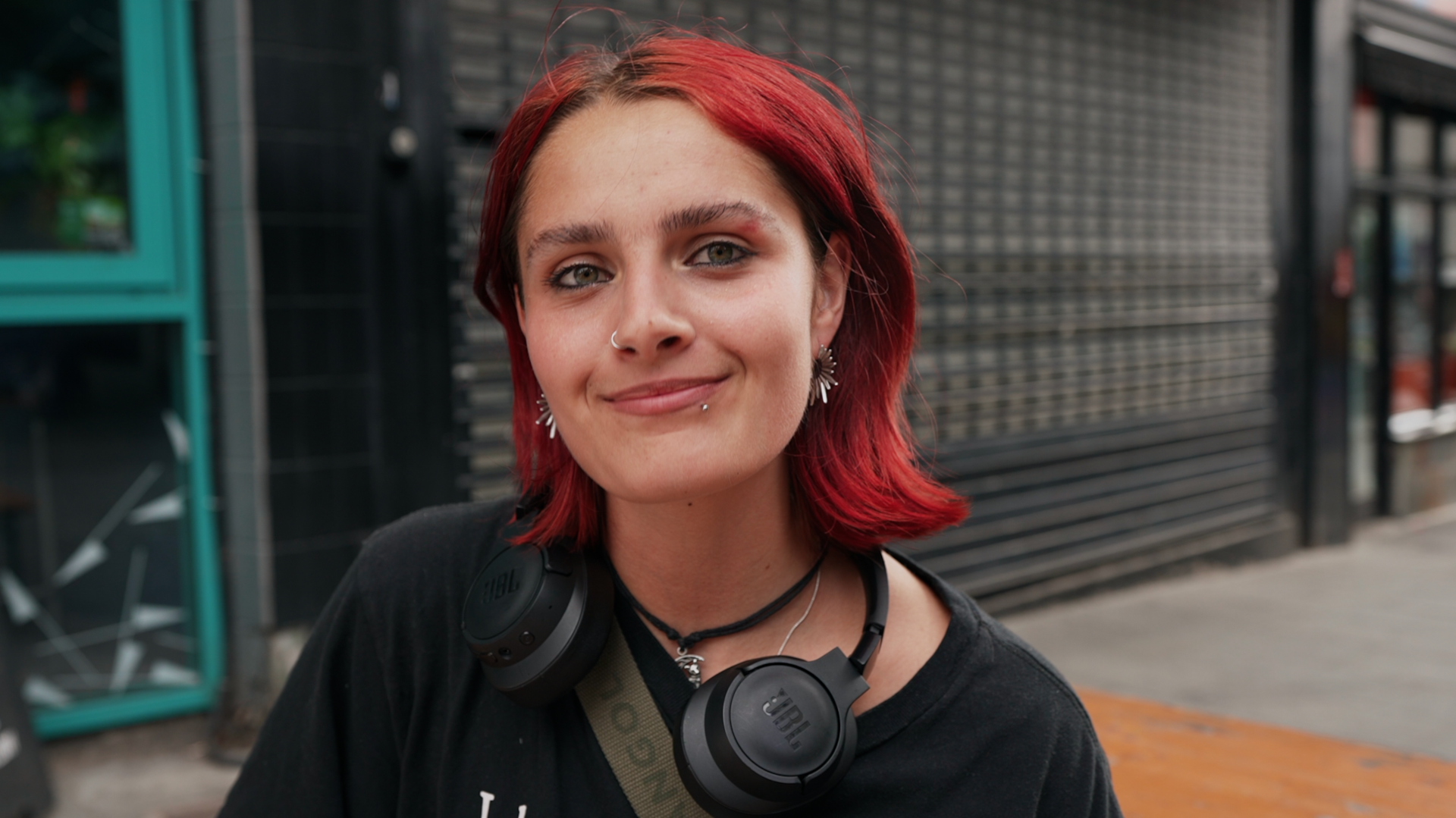 young woman with short red hair in the foreground, wearing a black T-shirt, black over-ear headphones around the neck, a cross-body strap, a nose ring and earrings. The person is facing the camera on a pavement outside a building. Behind the person is a dark tiled wall and metal security shutter, with a turquoise-framed doorway visible on the left and part of a street scene on the right. No bags, bins or markers are visible.