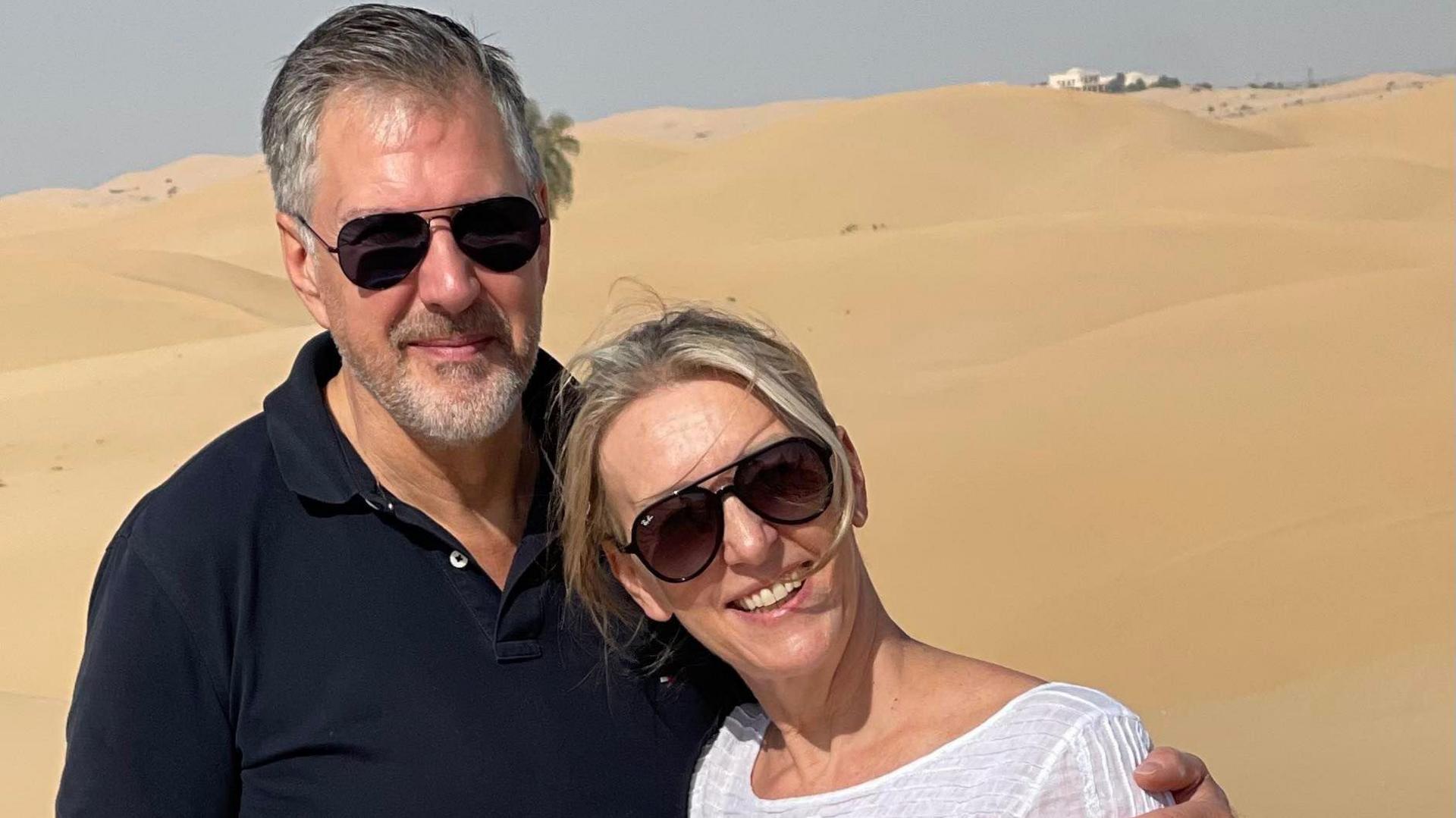 Angela Hudson, in a white top and aviator sunglasses, and her husband Chris, in a black polo shirt, with sand dunes behind them.