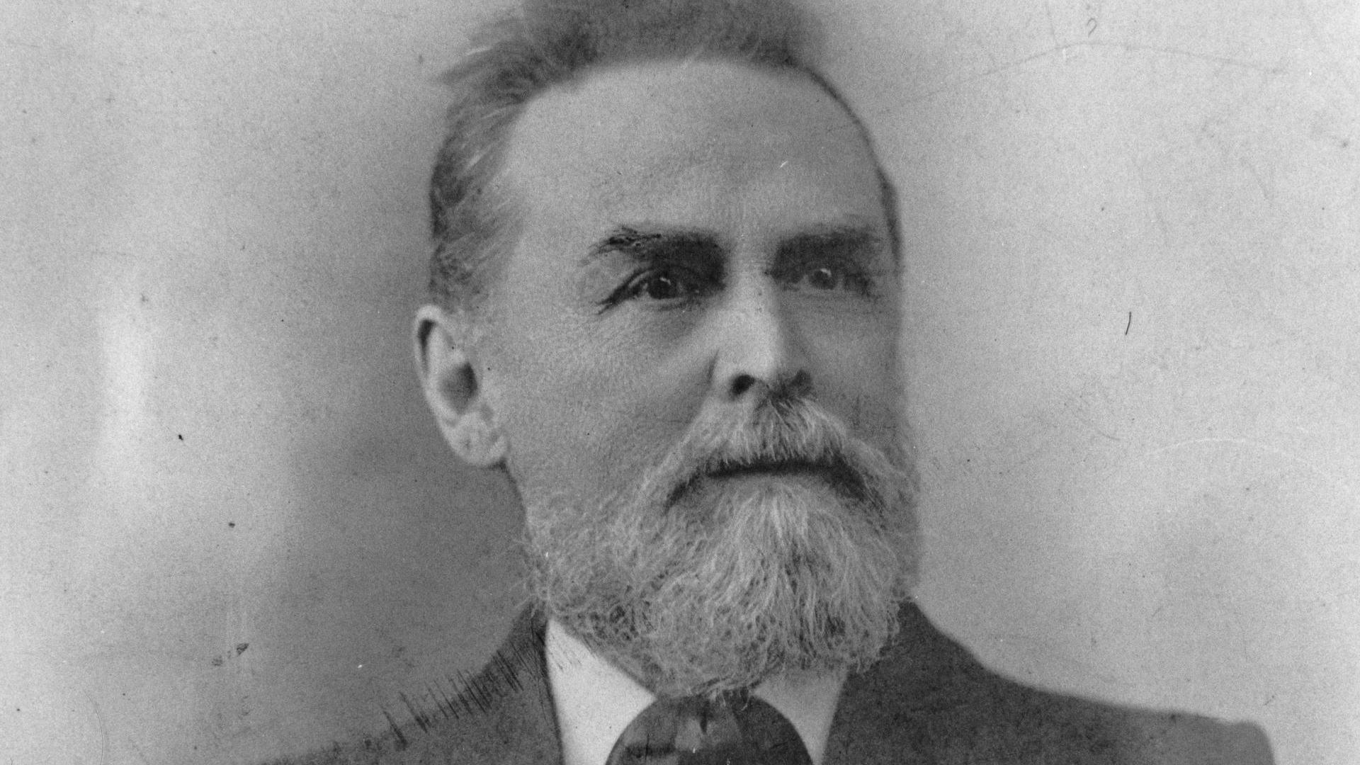 A black and white portrait photo of Frederick Hendy. He has a white beard and is wearing a black jacket, white shirt and dark tie. He is in front of a plain grey background, 