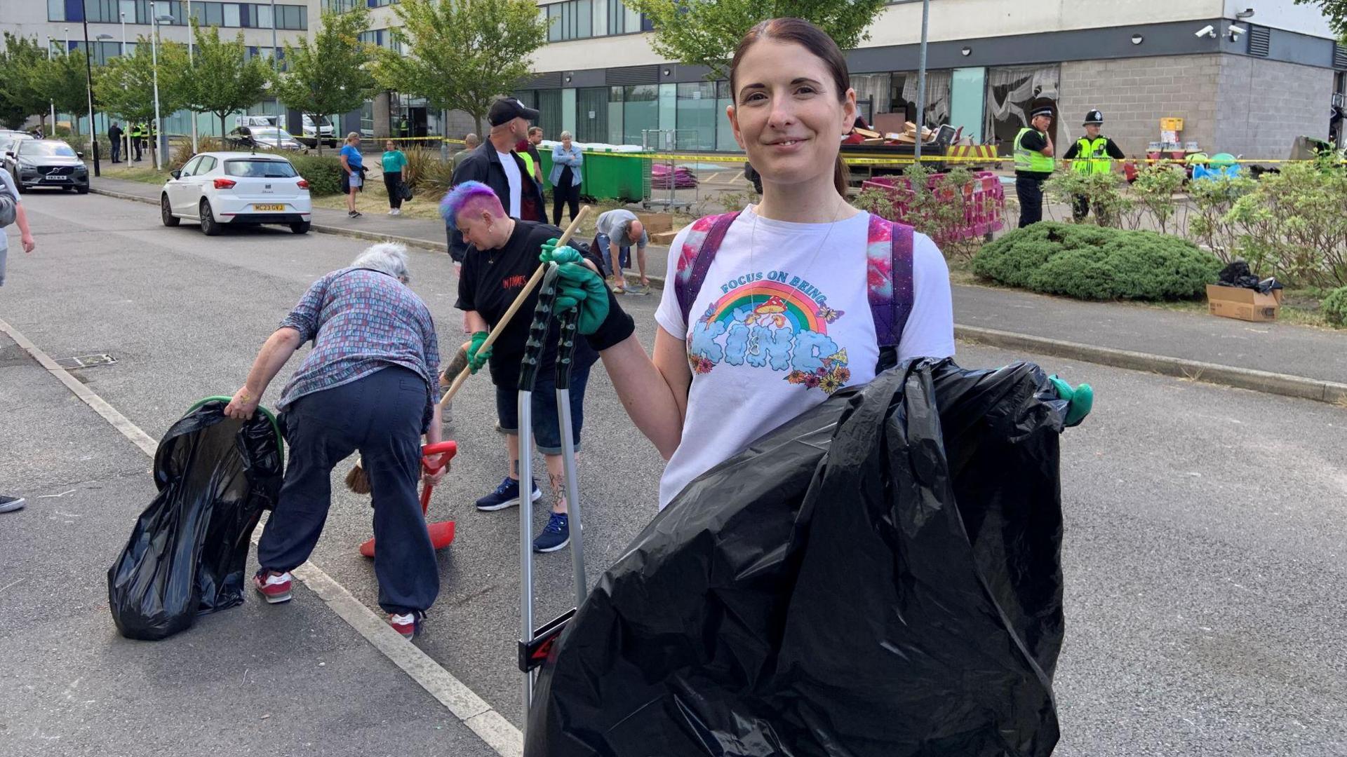 A woman stands in the middle of the picture, holding a rubbish bag and a litter picker. 