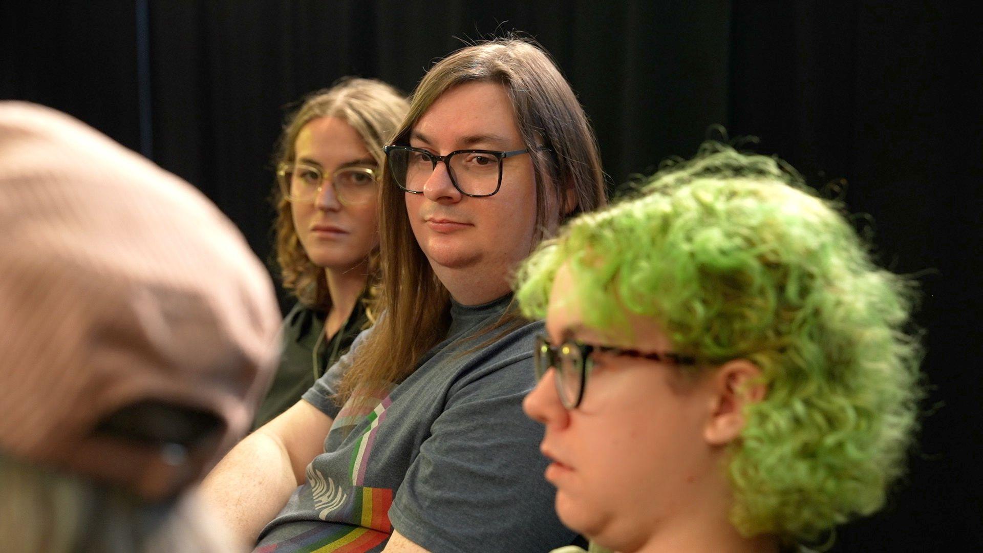 An candid image showing three people in a focus group listening while another person off-camera is speaking. Benji Rayson is in the middle of the image, looking towards the left of the frame. They are wearing a grey t-shirt and have straight brown hair that hangs below their shoulders. They have dark-rimmed rectangular glasses and have a neutral expression on their face.