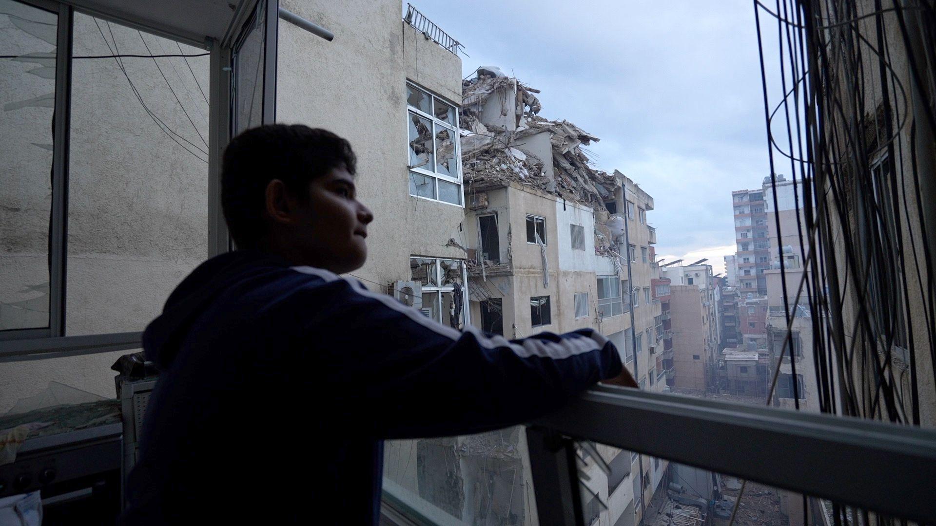 Boy looks out from a balcony, facing right. Damaged buildings can be seen.