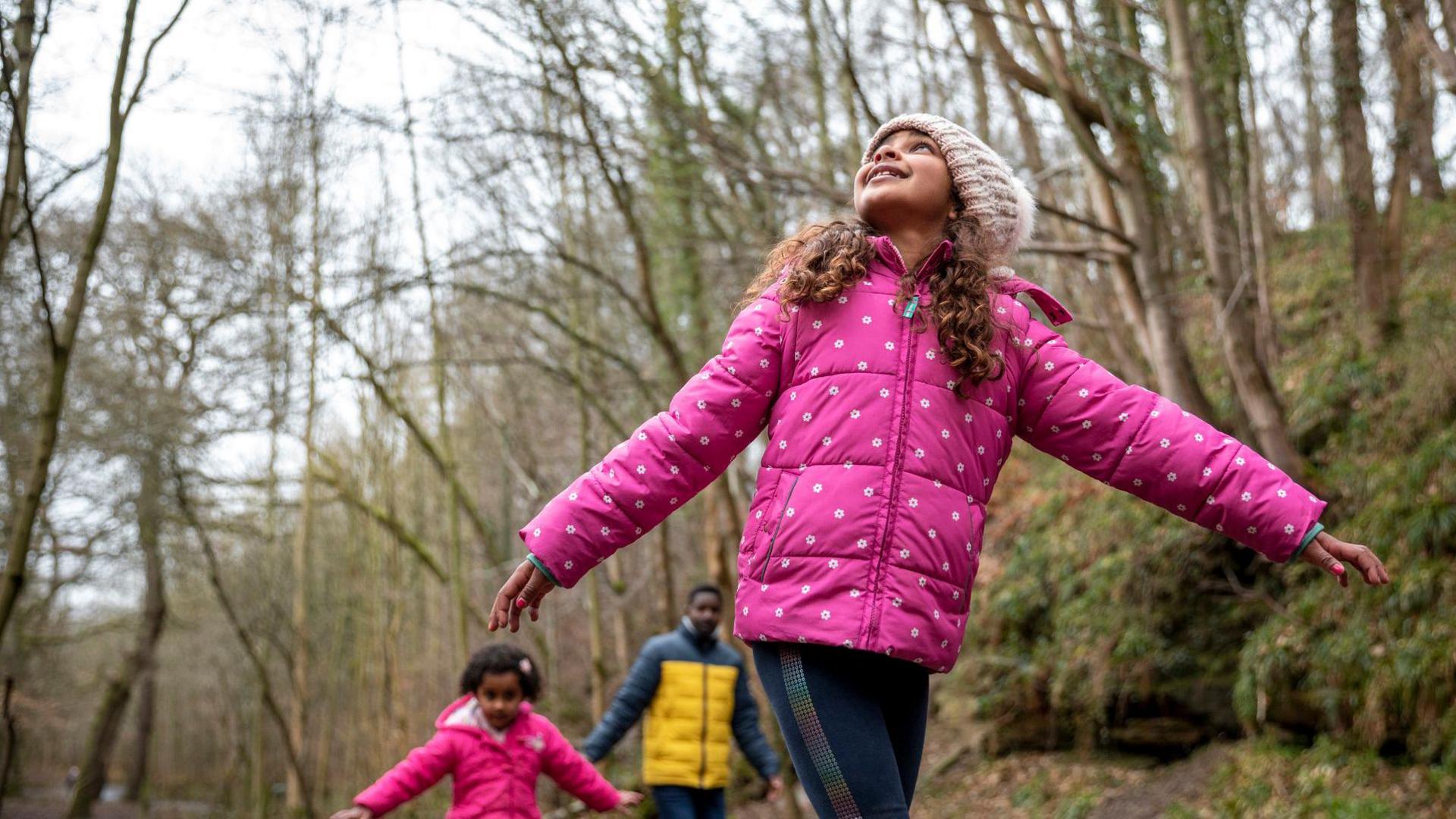 A child and their family walk in the woods, looking up at the tall trees.