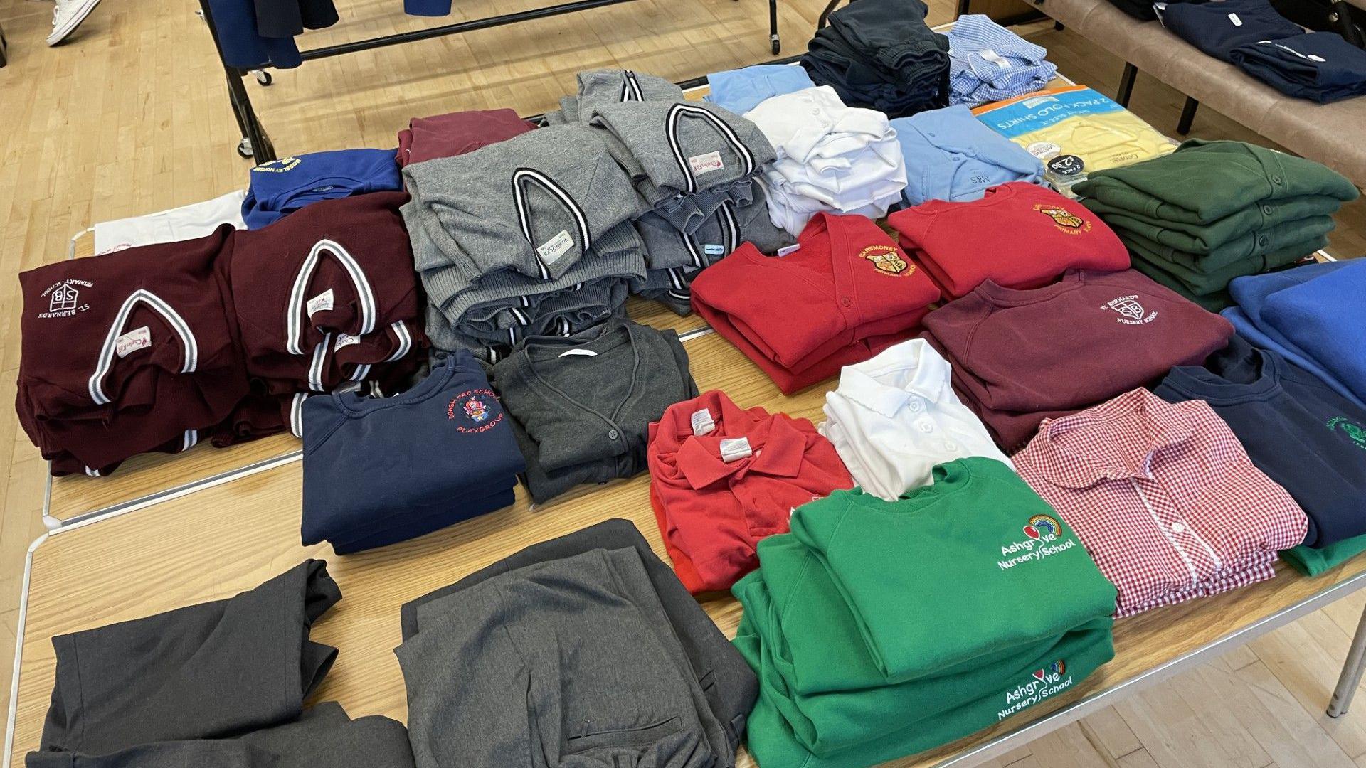 Piles of red, navy, grey and maroon uniform pieces are stacked on a brown table