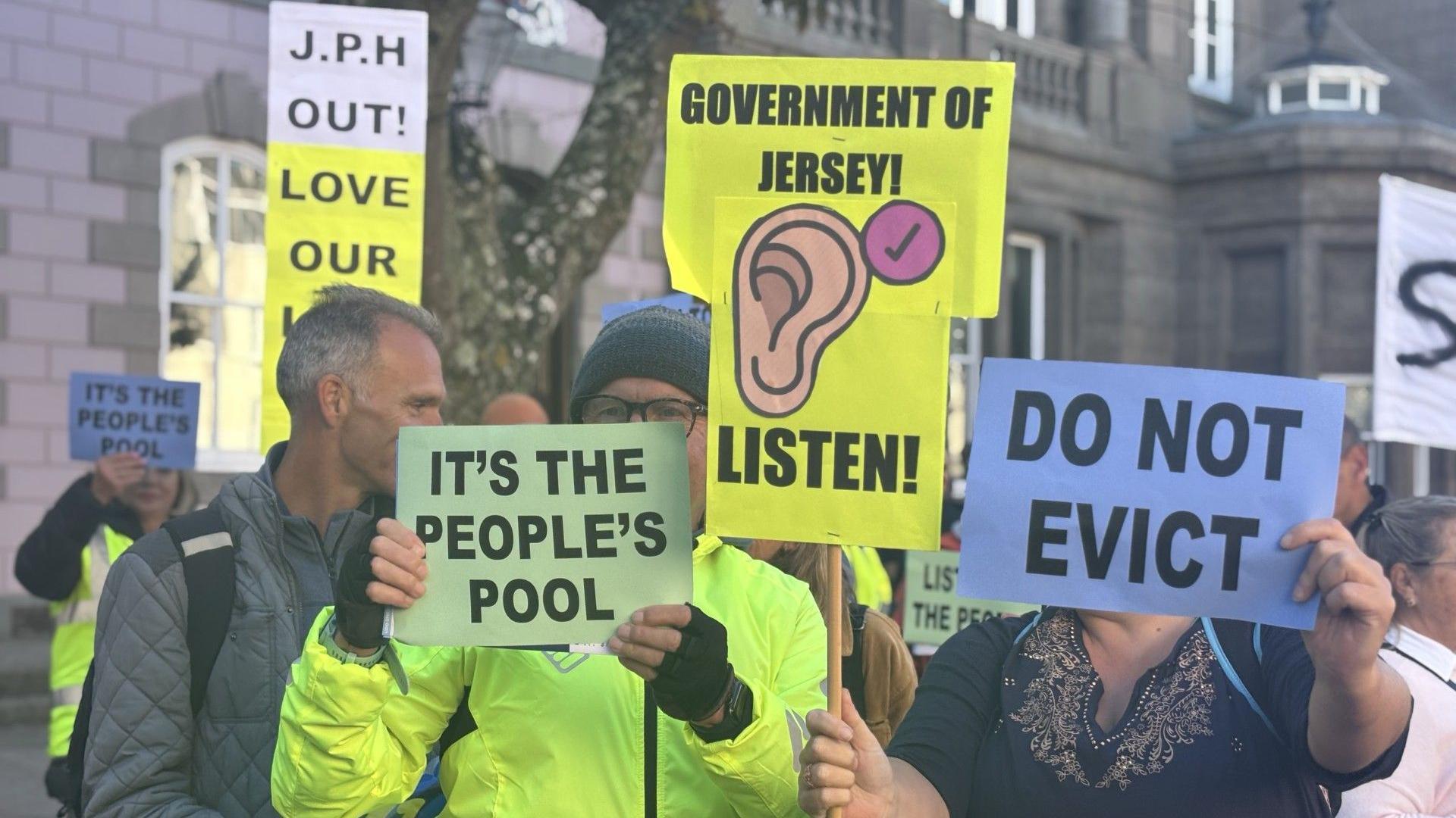 A man in a high vis green jacket is holding a sign saying "It's the people's pool" . Another placard has a ear on it and it says "Government of Jersey- Listen!" Another placard says "Do Not Evict". 