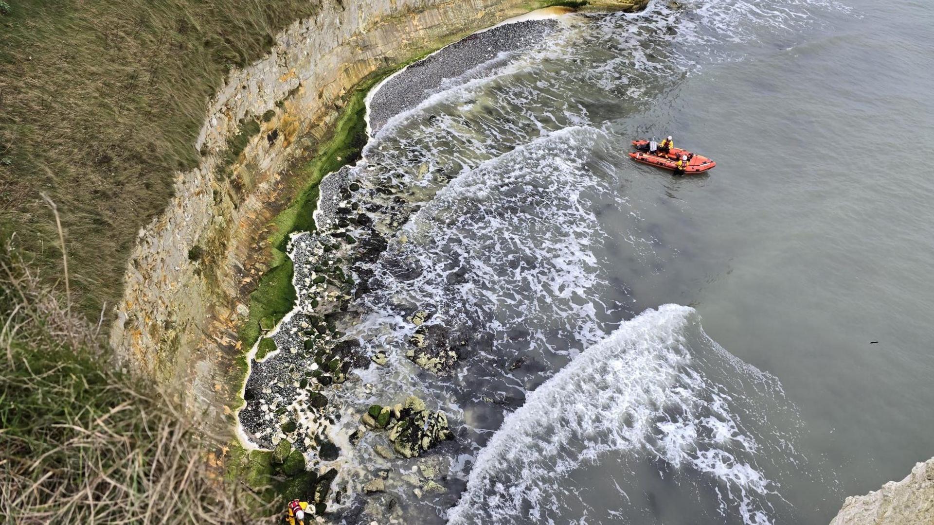 In this view a lifeboat is seen from above in the sea. A crewmember has reached the shore and is making their way to the dog.