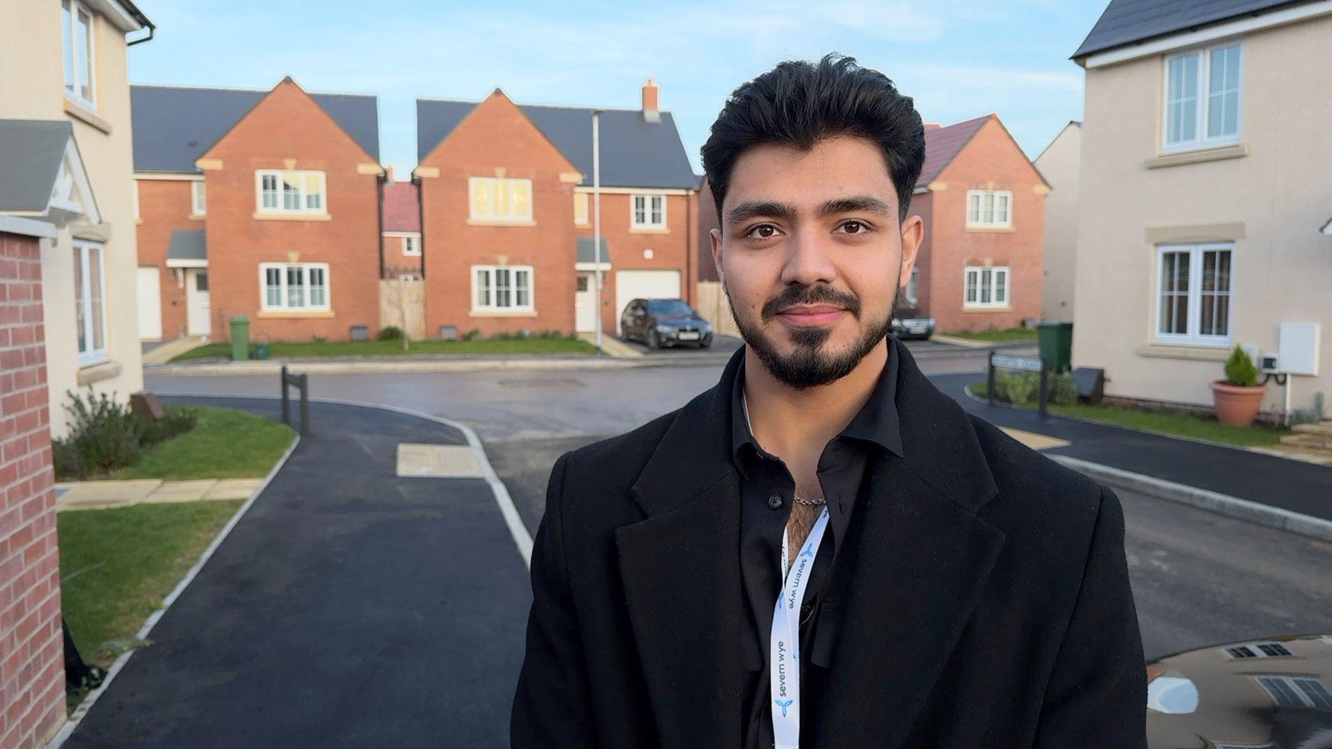 A man with short black hair, a beard and wearing a black shirt, jacket and lanyard, stands in the middle of a housing estate.