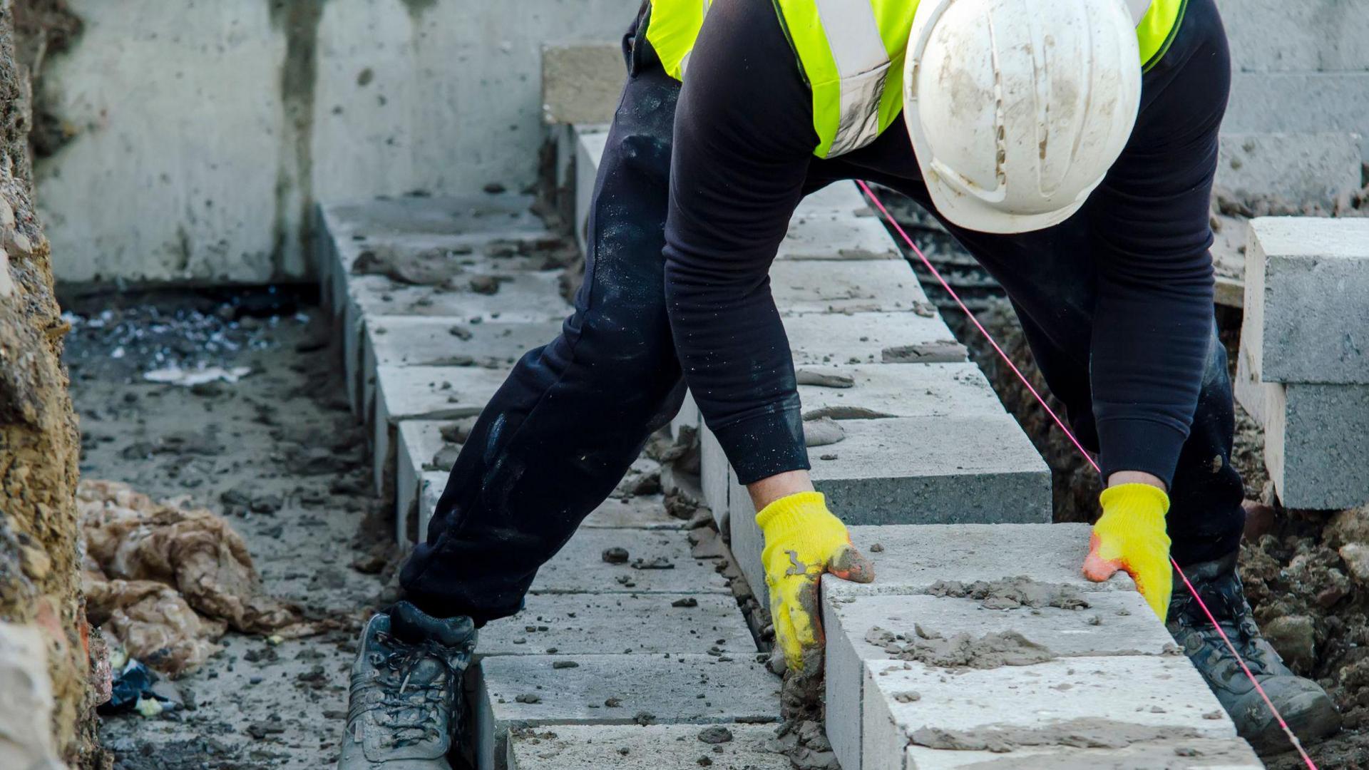 Construction worker wearing high-vis vest, white helmet and yellow gloves, bending over laying large grey bricks