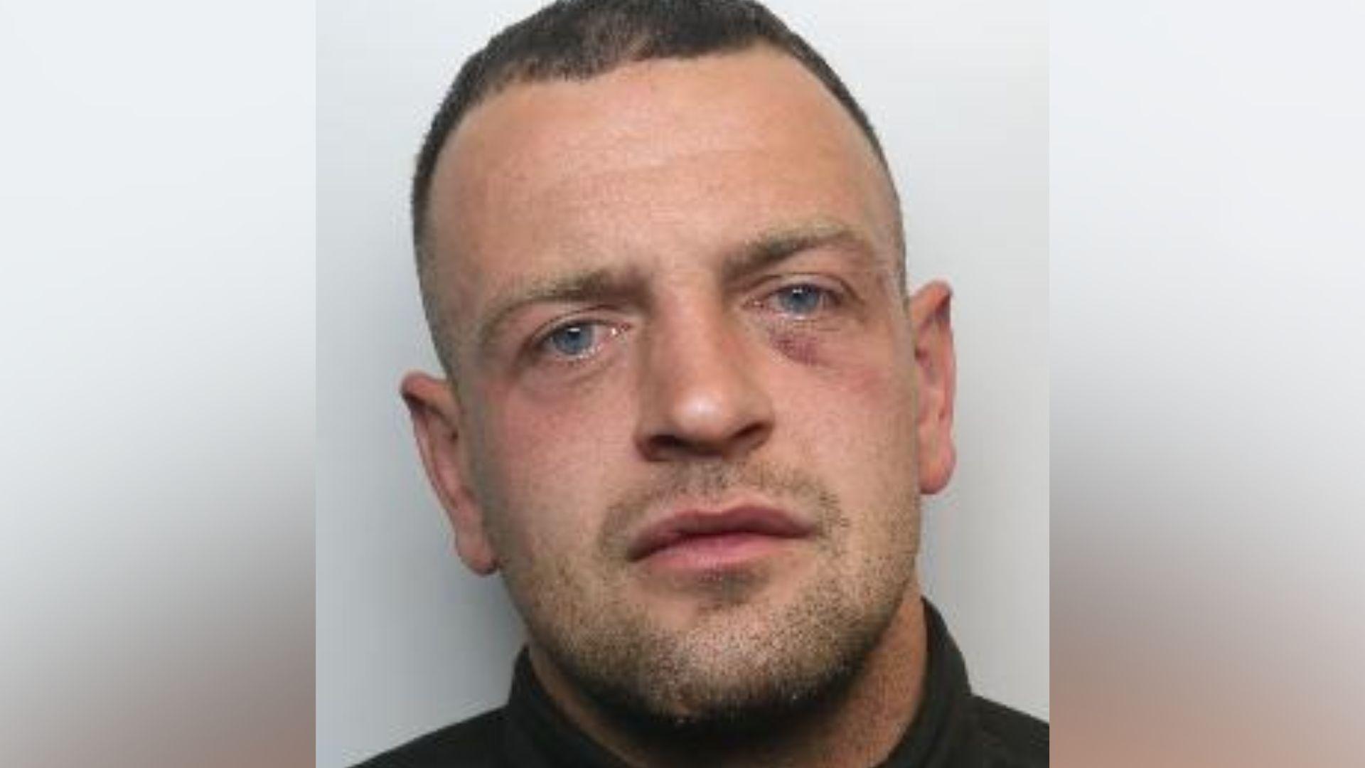 A police mugshot of a man wearing a dark-coloured top against a plain light background.
