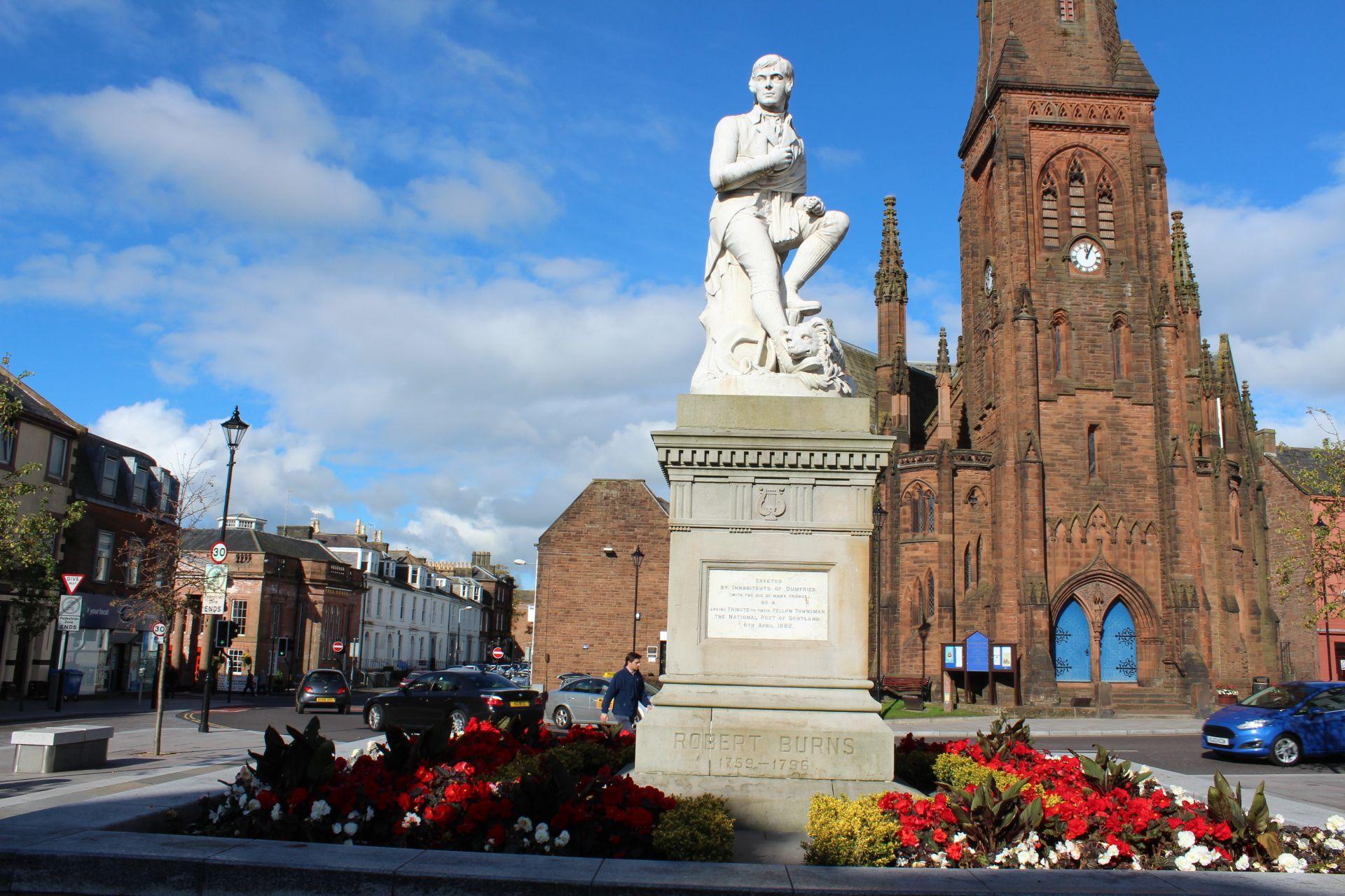 Burns Statue in Dumfries in front of Greyfriars Church - a large sandstone building - on a sunny day.