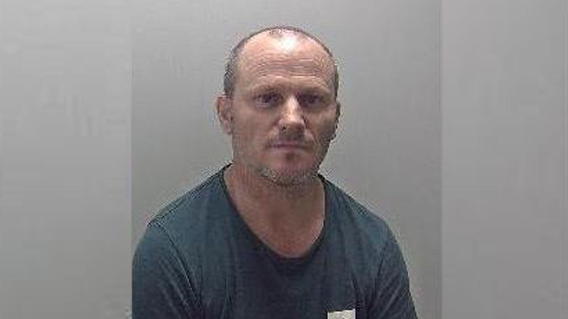 A mugshot of a man with receding brown hair and stubble, in a dark T-shirt 