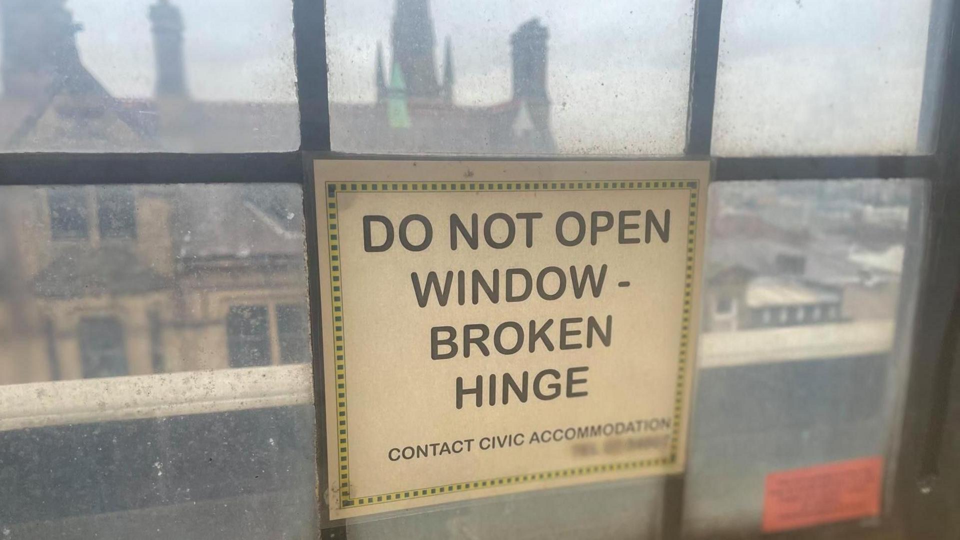 A smeared window looking out at an old historic building. A sign on the window reads, "Do not open, broken hinge"