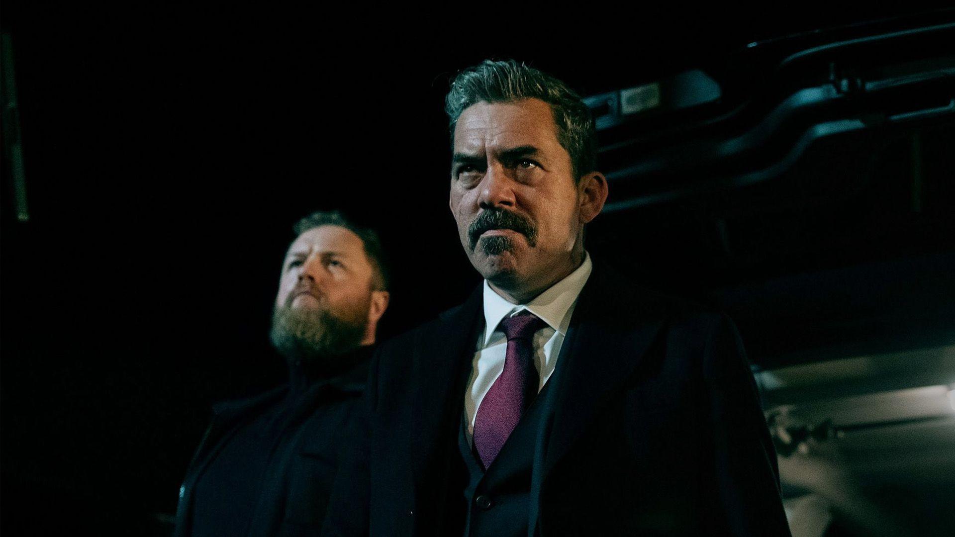 Two men are looking forward. It's dark.
The man in the foreground has greying short hair and a moustache.
He is wearing a dark coat, over a dark suit. His shirt is white and his tie purple.
The man behind is dressed fully in black. He has lighter hair and a full beard