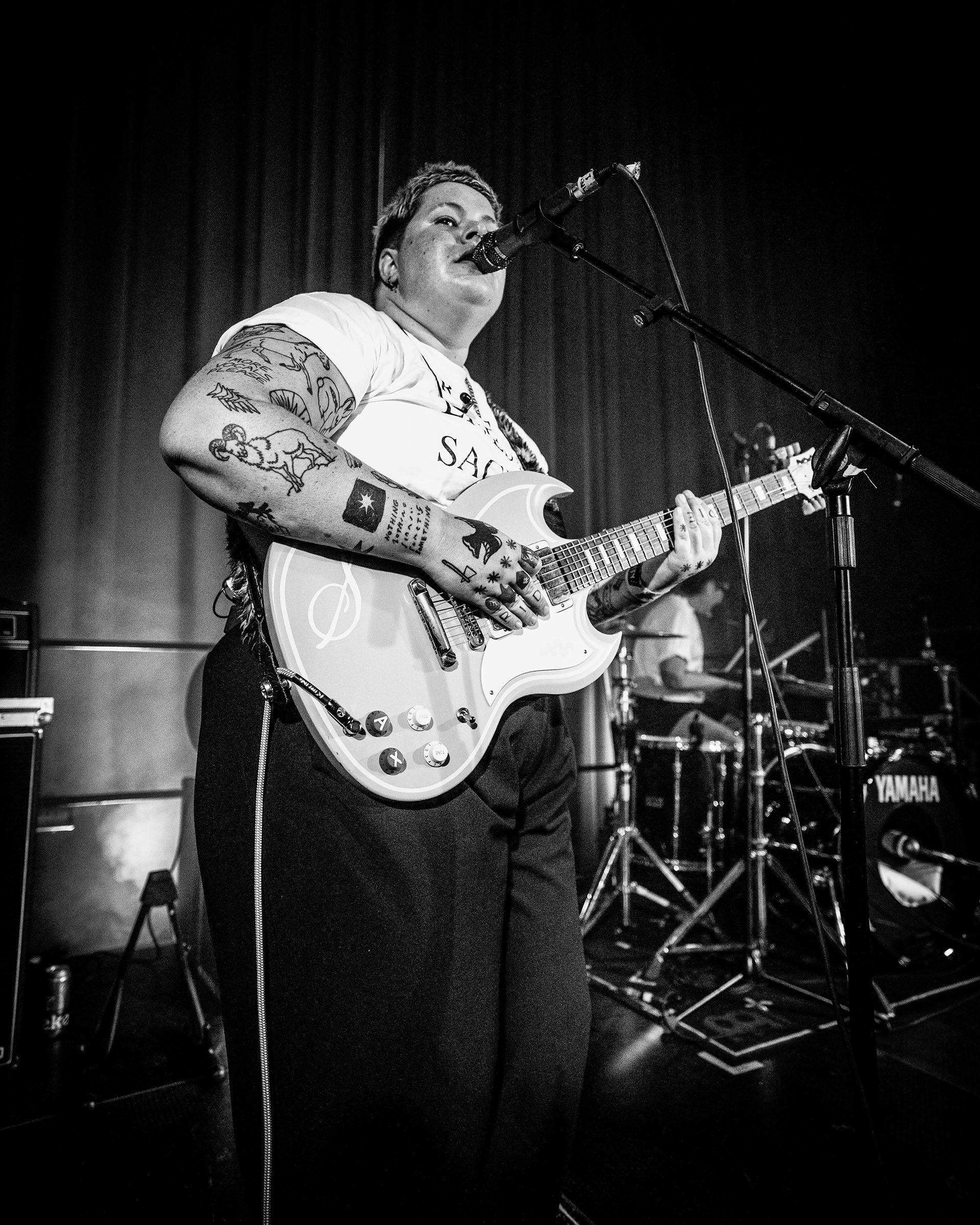 a black and white picture of hanni pidduck from the band arxx playing a gig. they are playing a guitar in front of a microphone. they are wearing a white t-shirt and dark trousers
