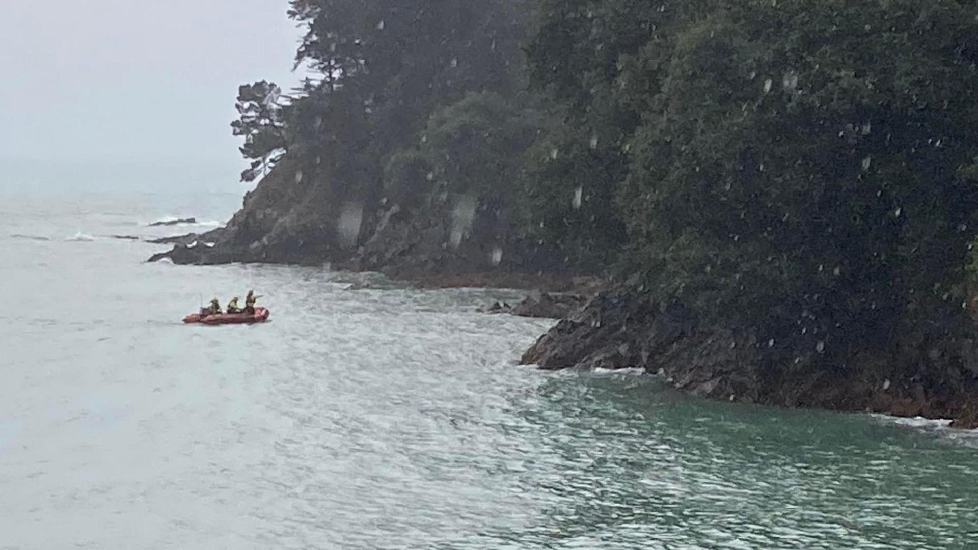 A rescue team on a boat on the water heading to a set of coastal rocks where two swimmers are waiting to be retrieved on a rainy day.