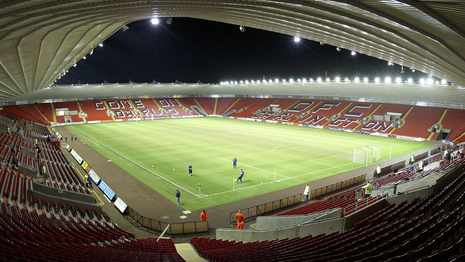 A flood-lit 25,000 seat football stadium with players training on the pitch, viewed from the back of the stands through a len that takes in the whole panorama of seats, green pitch and encouraging messages written across the rows of seats.