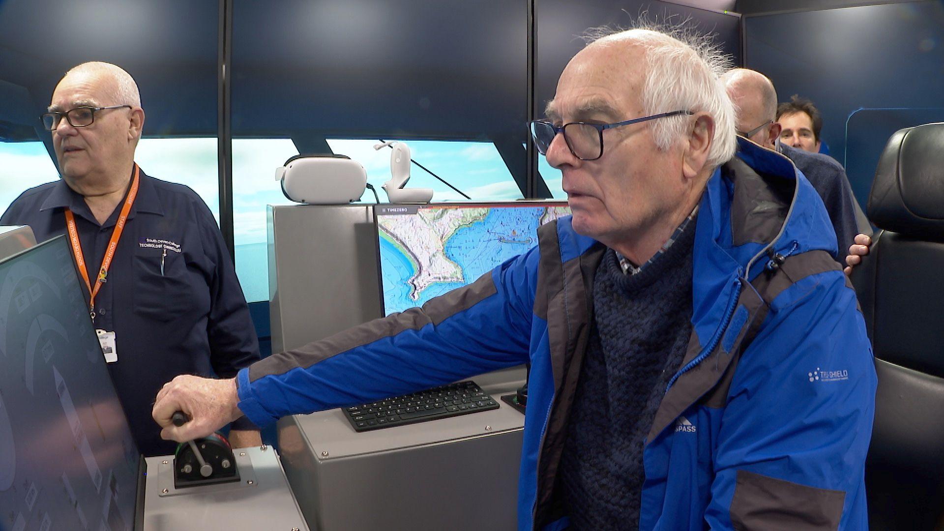 Retired fishermen Colin Warwick trying to steer a boat in the navigational simulator at the Marine Academy in Kingswear. He is wearing a blue and black coat and has white hair and black glasses. Fishing maps are behind him.