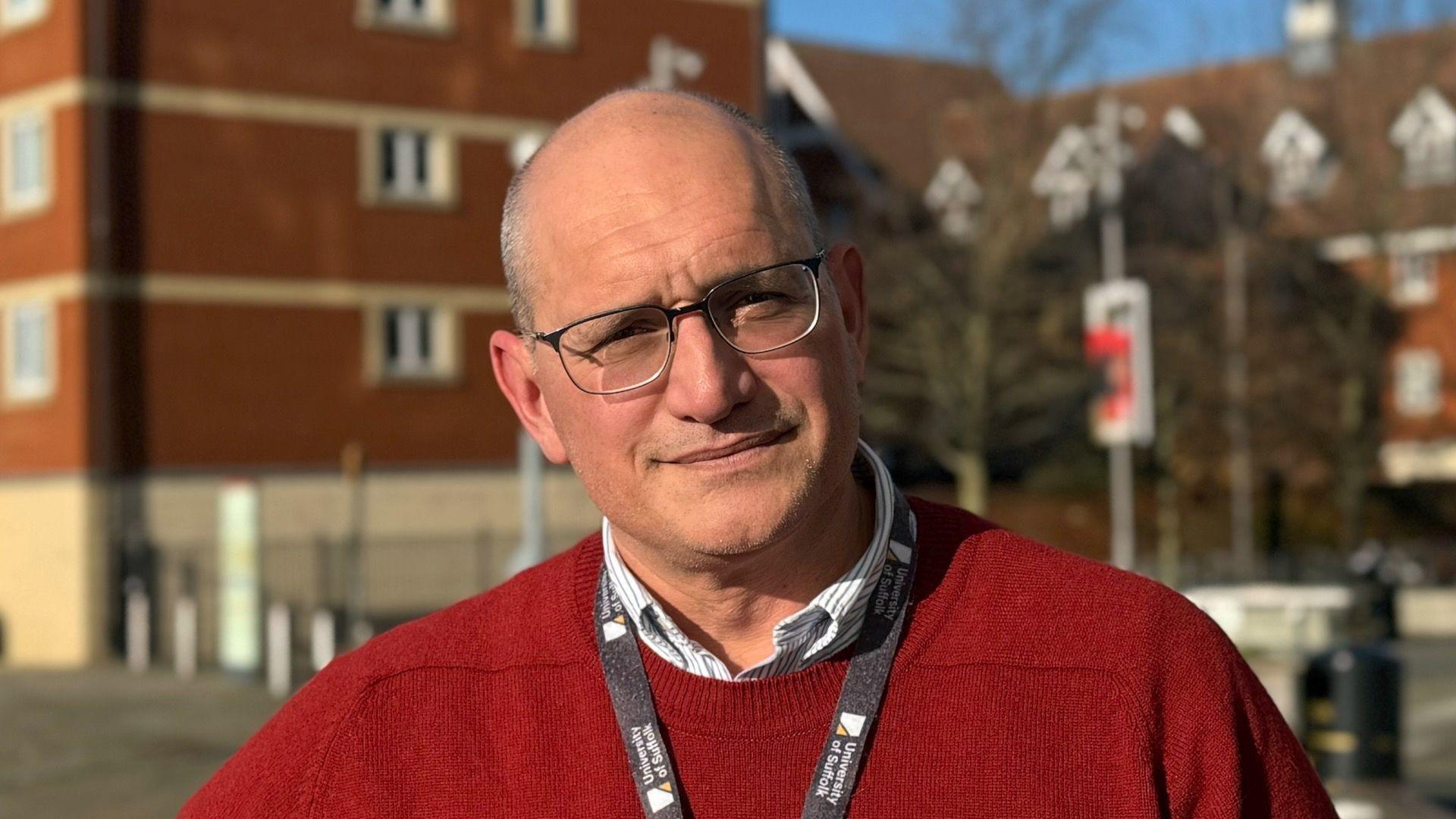 A man - Mike Sauter - looking straight at the camera. He is wearing a red jumper over a white shirt and has a black University of Suffolk lanyard around his neck.