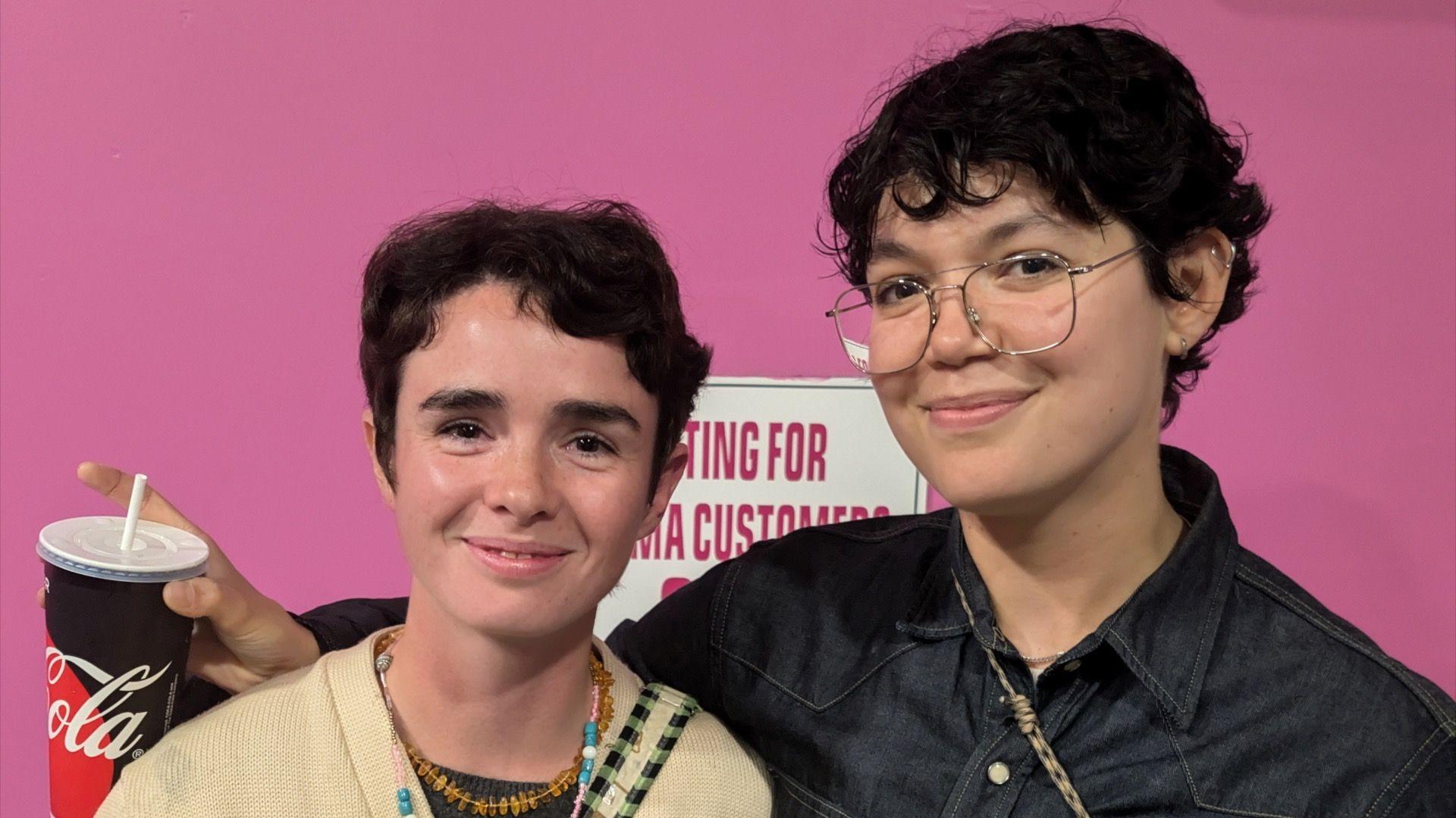 Filmmaker Iso, left, and artist Mol side by side in front of a pink background. Both have short dark hair and are smiling at the camera. Mol also has silver-rimmed glasses.