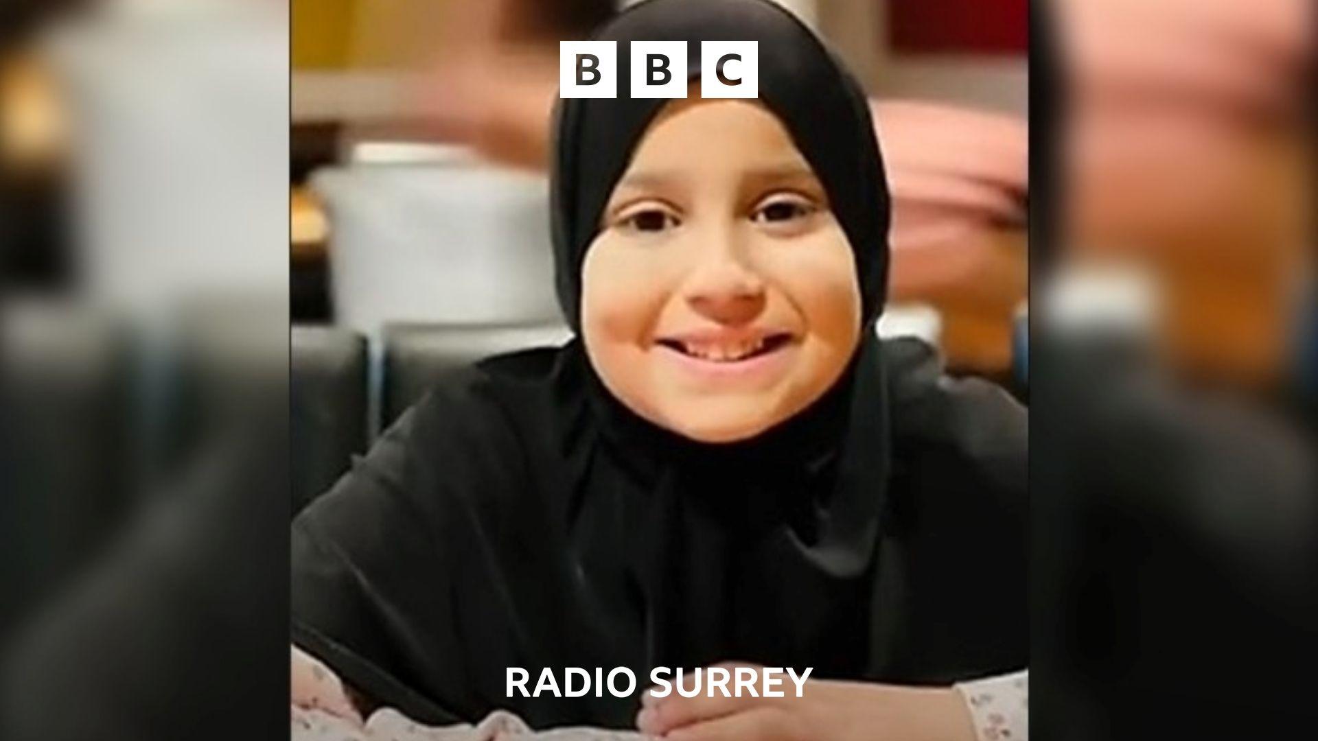 A photo of Sara Sharif. She has her hands on the table in front of her and is smiling at the camera. She is wearing a black hijab.