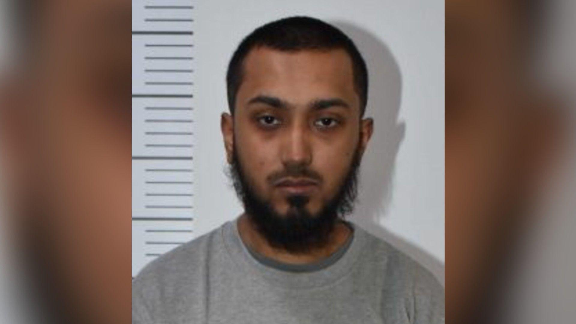 A custody image of Mohammed Mahfuz Ahmed. He is standing against a white wall with a height chart and is wearing a grey sweatshirt. He has short dark hair and a thick beard.