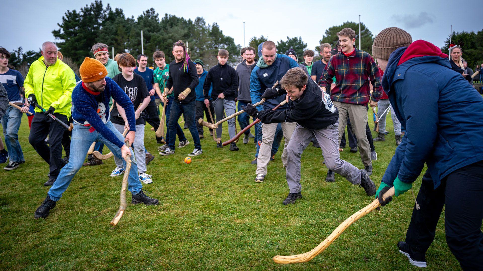 People in winter jumpers and hats grapple to hit a small orange ball with homemade sticks.