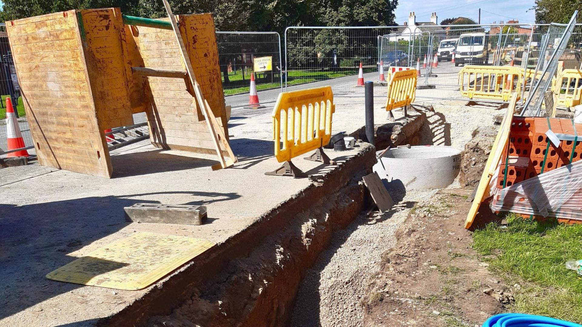 A building site, with pipes being laid alongside a road. There are yellow plastic barriers and metal fences surrounding the works.