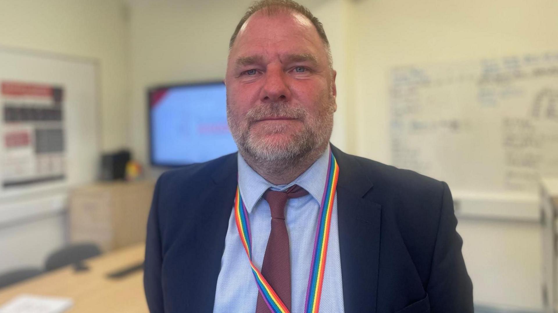 A man in a navy suit jacket and tie with a grey beard smiles for the camera