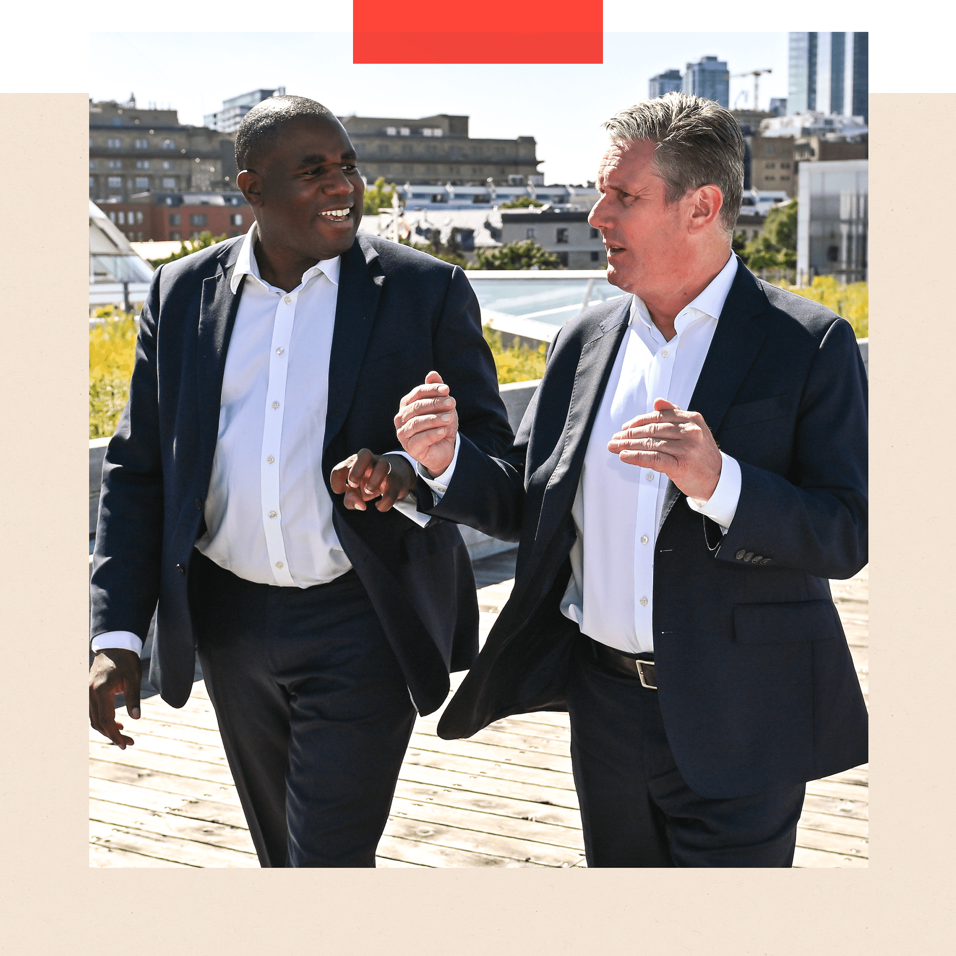 David Lammy on the left of shot and Keir Starmer on the right of shot. The men are outside and both wearing a dark suit and white shirt with no tie. Starmer has his arms out as though he is gesticulating and Lammy is looking at him and smiling