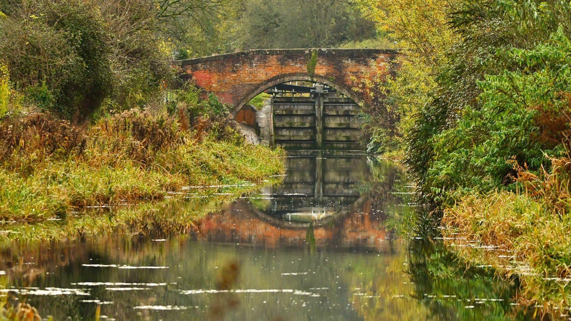 A bridge reflected in water below it, with greenery all around and a wooden lock in the background