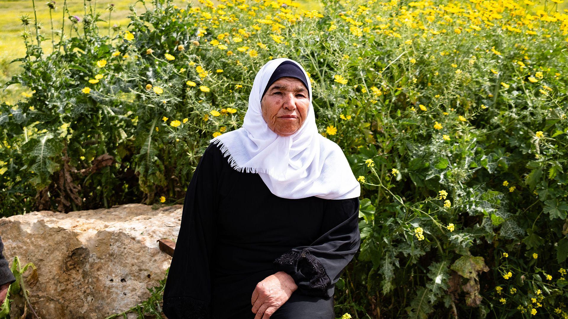 Palestinian grandmother Ayesha Shtayyeh, wearing a long-sleeved black tunic and white headscarf, with a serious expression on her face. She  sits in front of a huge swathe of yellow flowers. 