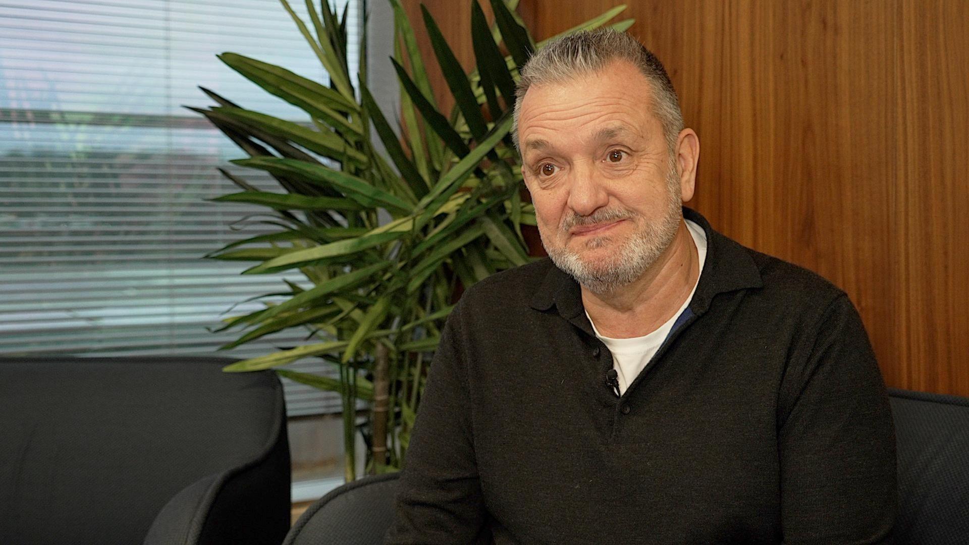 Gary Sweet is dressed in a black polo top and sat in a wood panelled room near window and potted plant.