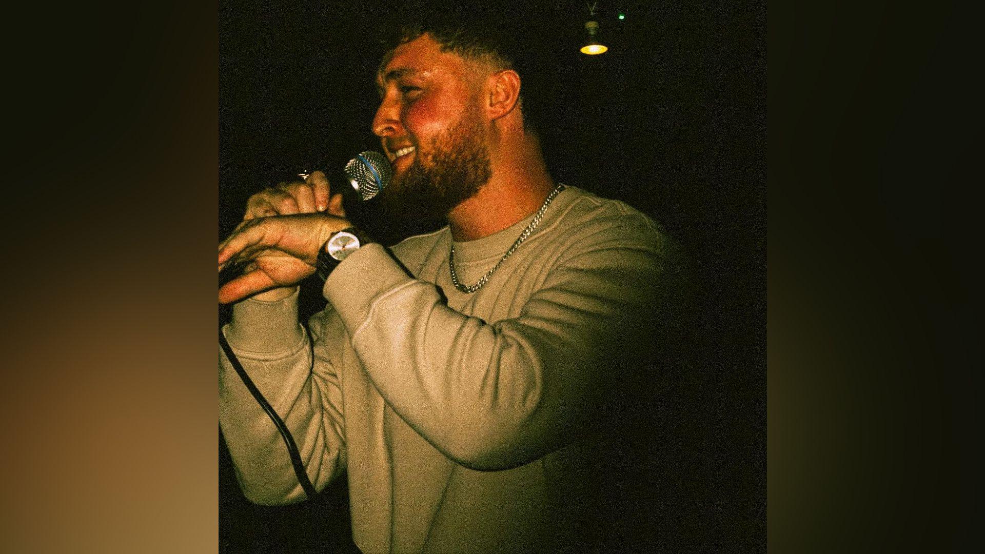 Ryan Finan is singing and smiling in a dark room under a golden light. He has brown hair and beard He is holding a microphone to his mouth.